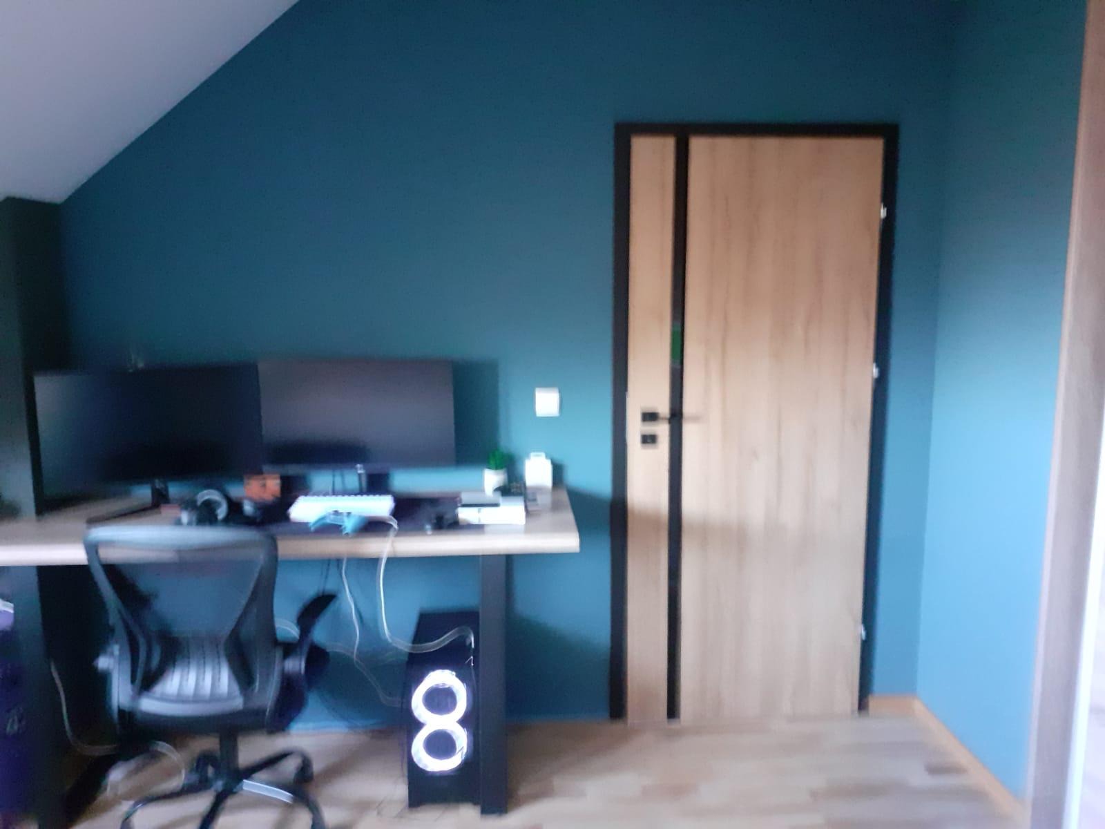 Pokój z biurkiem, dwoma monitorami, krzesłem obrotowym i komputerem z podświetleniem LED na tle ściany pomalowanej na kolor morski i drewnianych drzwi.