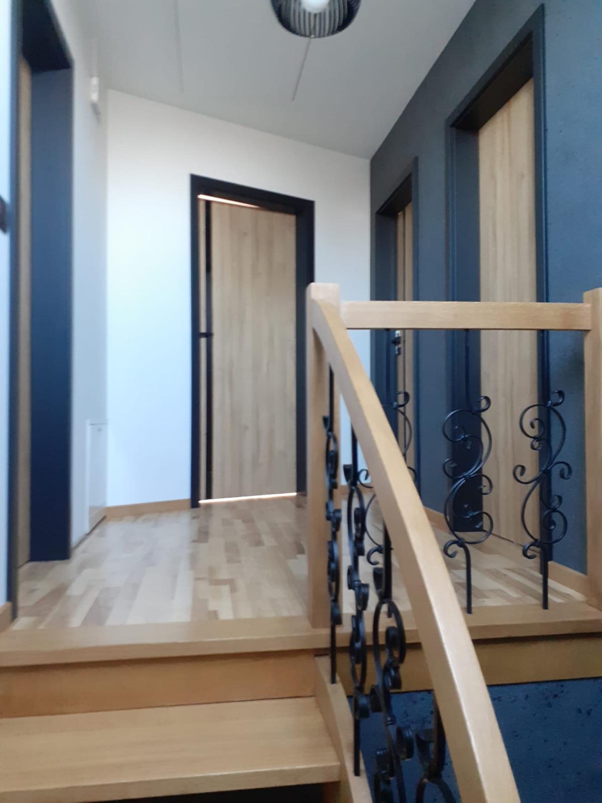 Korytarz z drewnianą podłogą, dwiema parami drzwi z czarnymi ościeżnicami i drewnianymi schodami z ozdobną, kutą balustradą.