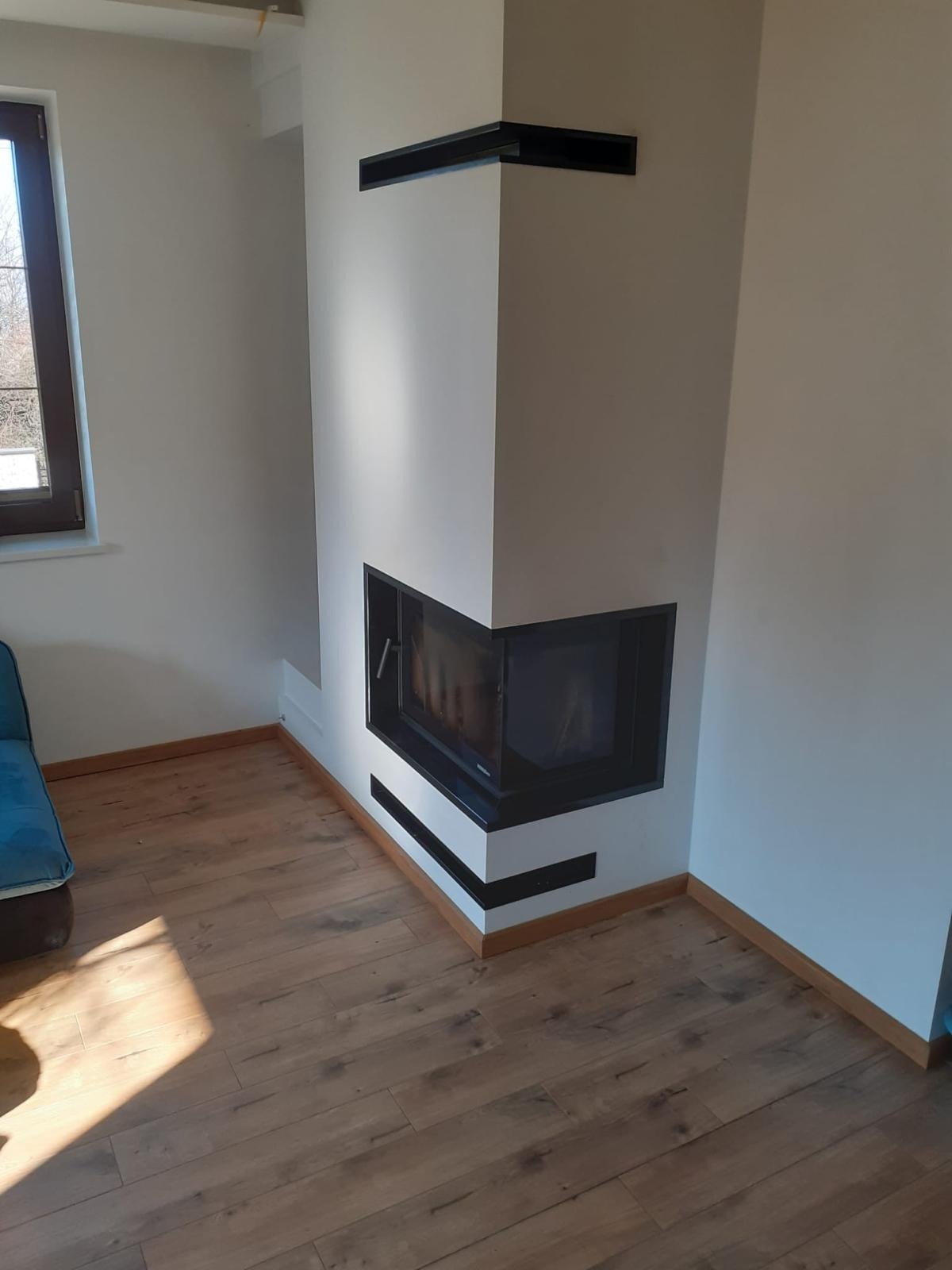 Nowoczesny narożny kominek z czarną obudową, w jasnym wnętrzu z drewnianą podłogą i fragmentem niebieskiej sofy.