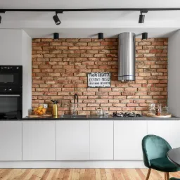 Nowoczesna kuchnia z białymi szafkami, ceglaną ścianą i czarnymi reflektorami sufitowymi, uzupełniona o zielone krzesło i drewnianą podłogę.