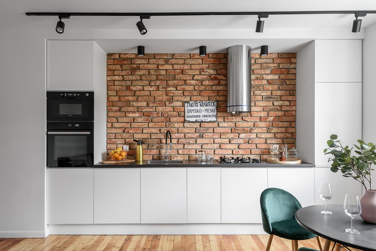 Nowoczesna kuchnia z białymi szafkami, ceglaną ścianą i czarnymi reflektorami sufitowymi, uzupełniona o zielone krzesło i drewnianą podłogę.