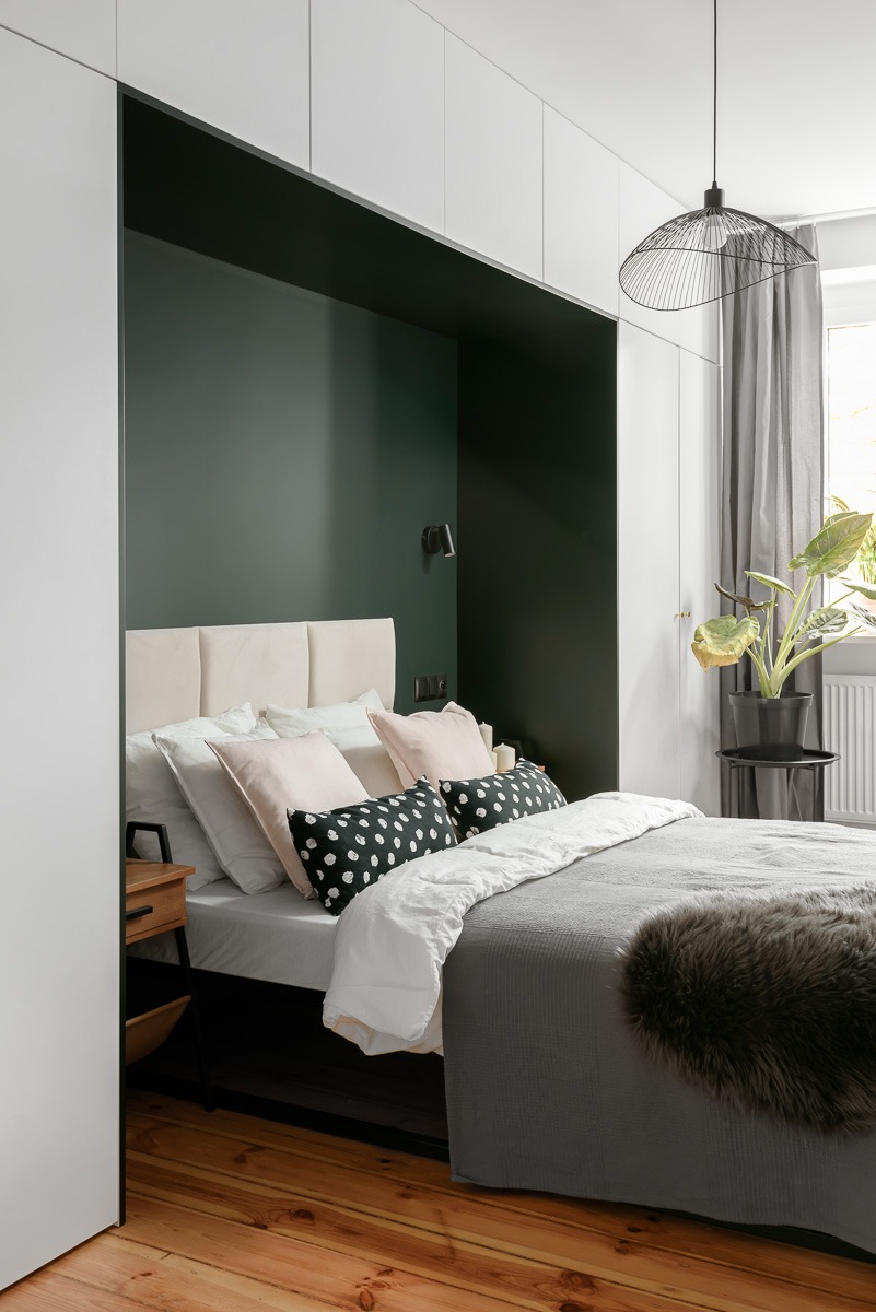 Nowoczesna sypialnia z wnęką w kolorze butelkowej zieleni, beżowym zagłówkiem i drewnianą podłogą. Czarna lampa druciana zwisa nad łóżkiem z szarą narzutą i dekoracyjnymi poduszkami.