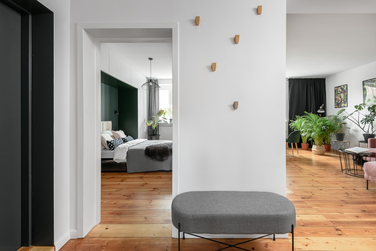 Wnętrze mieszkania z widokiem na sypialnię i salon, drewniana podłoga, szara pufa, biała ściana z drewnianymi wieszakami, czarne zasłony i rośliny doniczkowe w salonie.