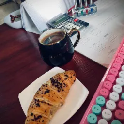 Przerwa w pracy biurowej: filiżanka kawy z napisem 'you deserve a break', czekoladowy rogalik na białym talerzyku obok różowej klawiatury, kalendarz biurkowy, kalkulator i zestaw długopisów...