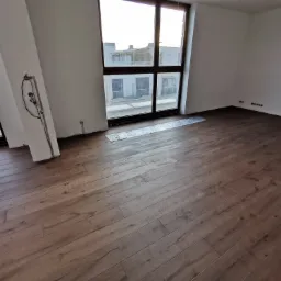 Nowo ułożona, brązowa podłoga z paneli w pustym pokoju z dużymi oknami i widocznymi instalacjami elektrycznymi na ścianach.