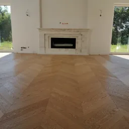 Jasna, drewniana podłoga ułożona we wzór jodełki francuskiej w nowym, przestronnym salonie z marmurowym kominkiem i dużymi oknami.