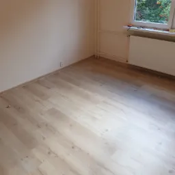 Pusty pokój po remoncie z panelami podłogowymi w odcieniach beżu i kremu, jasnymi ścianami i białym grzejnikiem pod oknem, na parapecie narzędzia i taśma malarska.