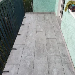 Wyłożony szarymi płytkami balkon z zieloną balustradą i ścianą o chropowatej strukturze, widok z góry na ułożone prostokątne płytki.