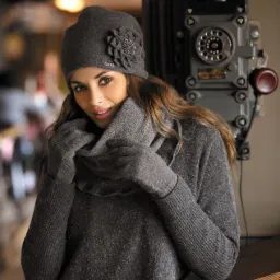 Portret kobiety w szarym komplecie: czapka z ozdobną aplikacją, rękawiczki i golf, pozującej przy zabytkowym telefonie.