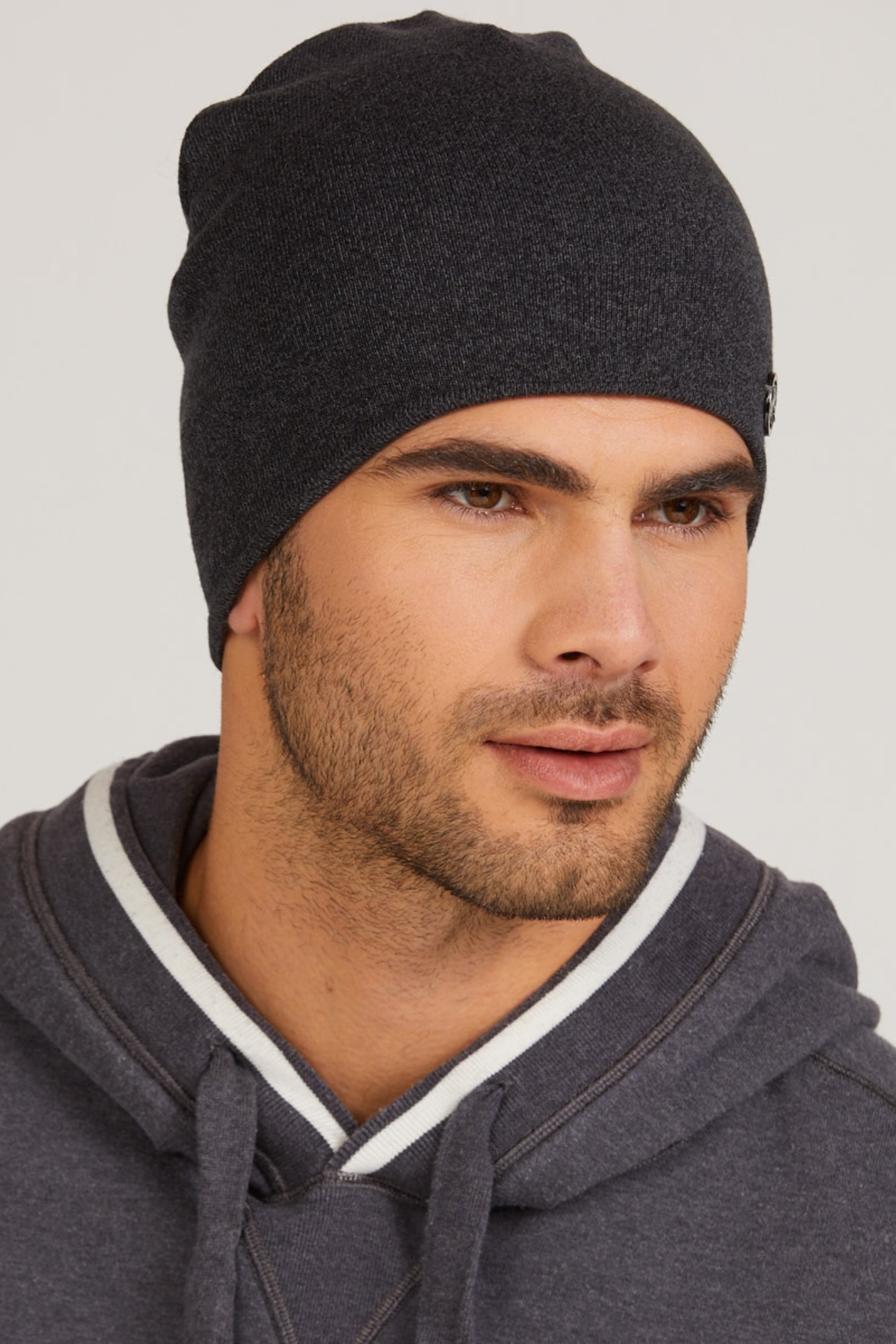 Mężczyzna w szarej czapce beanie z małym metalowym logo, ubrany w bluzę z kapturem z białym paskiem przy szyi, na jasnym tle.