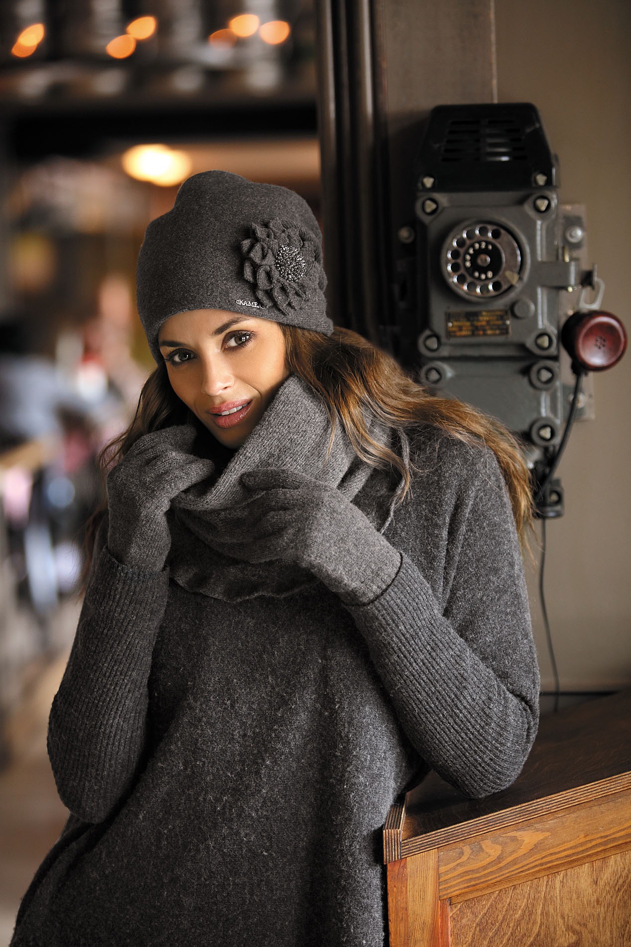 Portret kobiety w szarym komplecie: czapka z ozdobną aplikacją, rękawiczki i golf, pozującej przy zabytkowym telefonie.