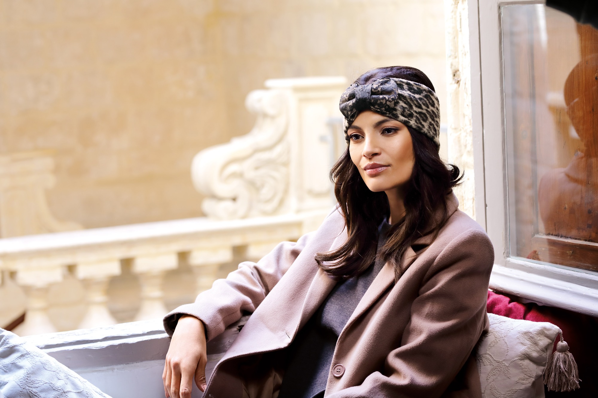 Kobieta w beżowym płaszczu i opasce w panterkę, siedząca na balkonie z kamienną balustradą, z widokiem na budynek w tle.