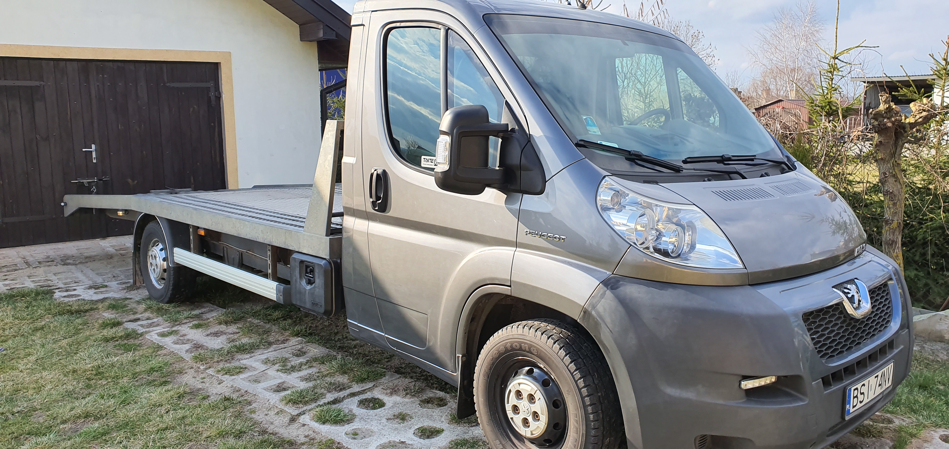 Szara laweta Peugeot zaparkowana na posesji z kostki brukowej i trawy, widoczna brama garażowa w tle.