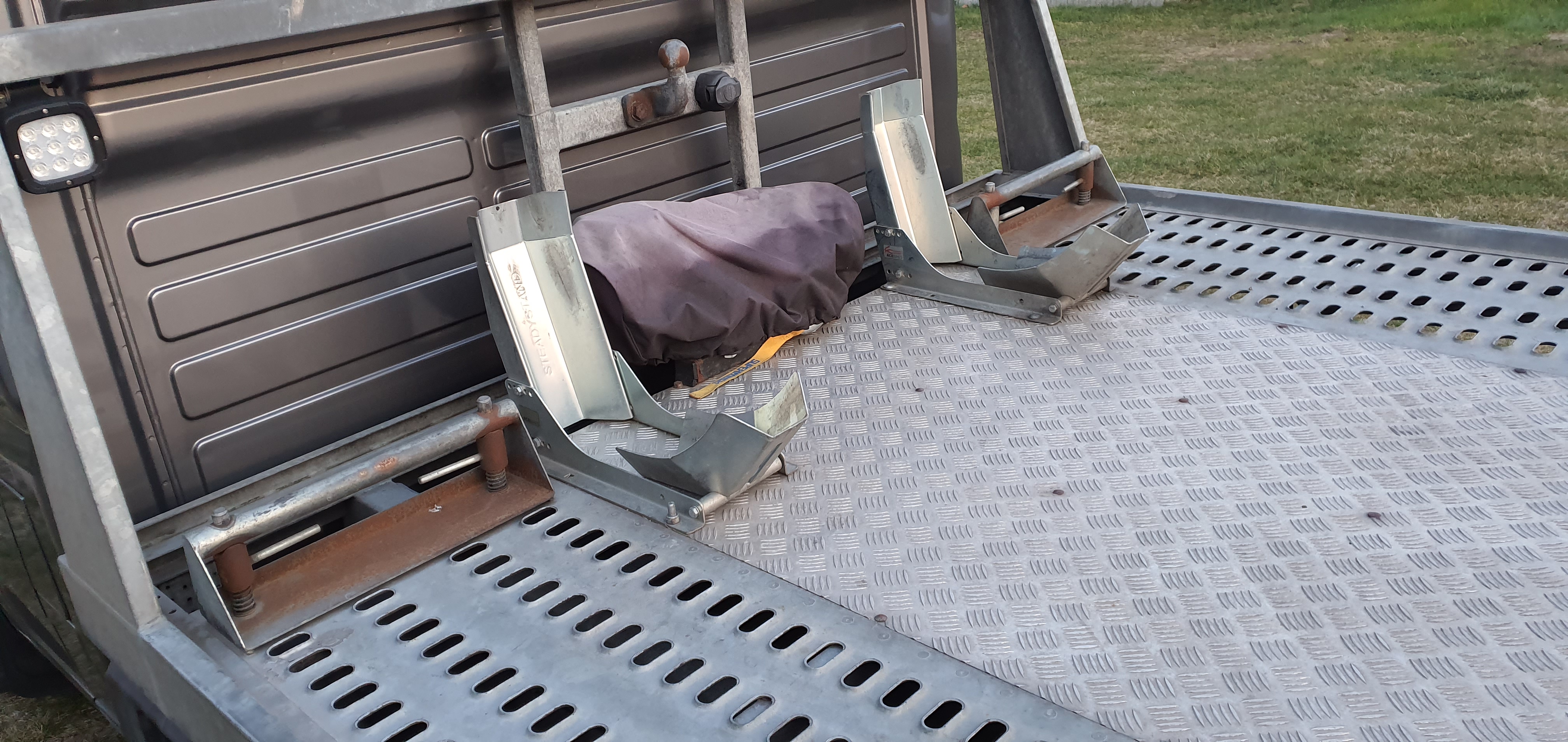 Pusta platforma lawety z metalowymi uchwytami na koła motocykla, zabezpieczona szarą plandeką, oświetlona lampą LED, widok z góry.