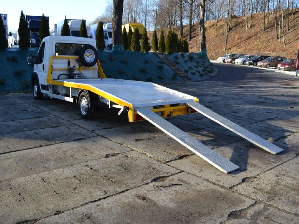 Biała laweta z żółtymi elementami konstrukcyjnymi, z opuszczonymi najazdami, zaparkowana na betonowym placu w słoneczny dzień.