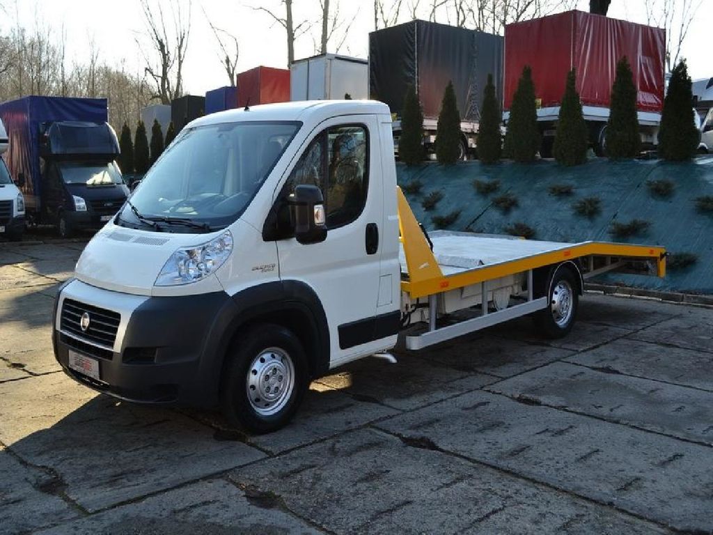 Biała laweta Fiat Ducato z żółtymi elementami platformy, zaparkowana na betonowym placu, z innymi samochodami dostawczymi i ciężarówkami w tle.