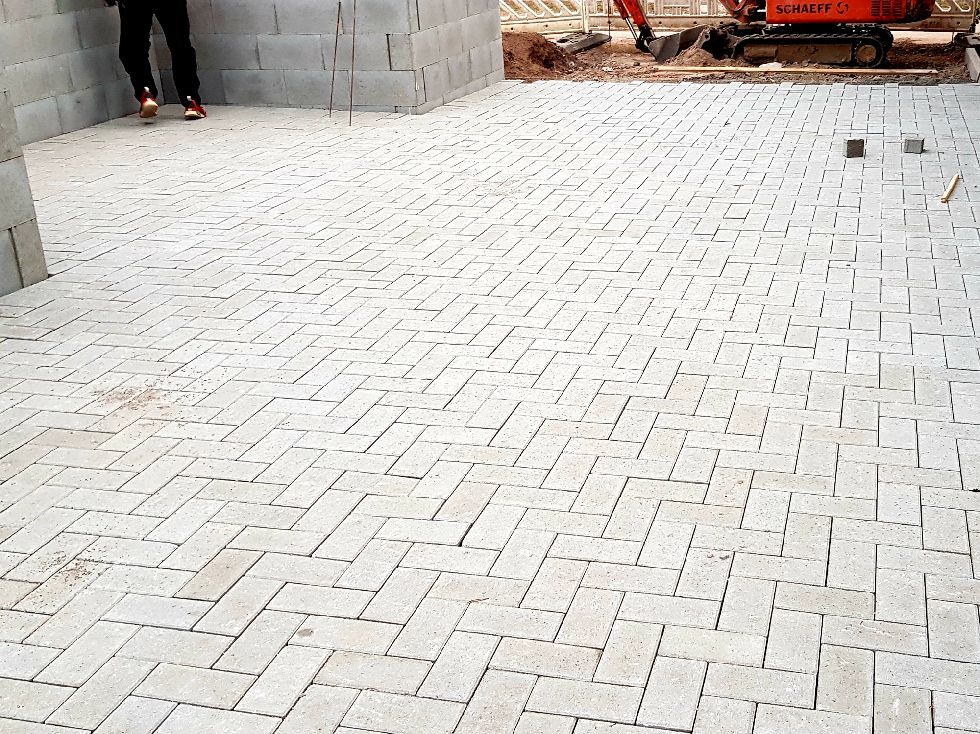 Ułożona jasnoszara kostka brukowa w jodełkę, z widocznym fragmentem fundamentu z bloczków betonowych i koparką w tle.
