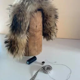 Wykończenie futrzane kołnierza na drewnianym manekinie krawieckim, obok igła, naparstek, nić i miarka krawiecka.