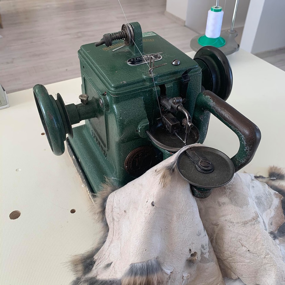 Stara, zielona maszyna szwalnicza szyje futro na jasnym stole; w tle widoczna szpula nici.