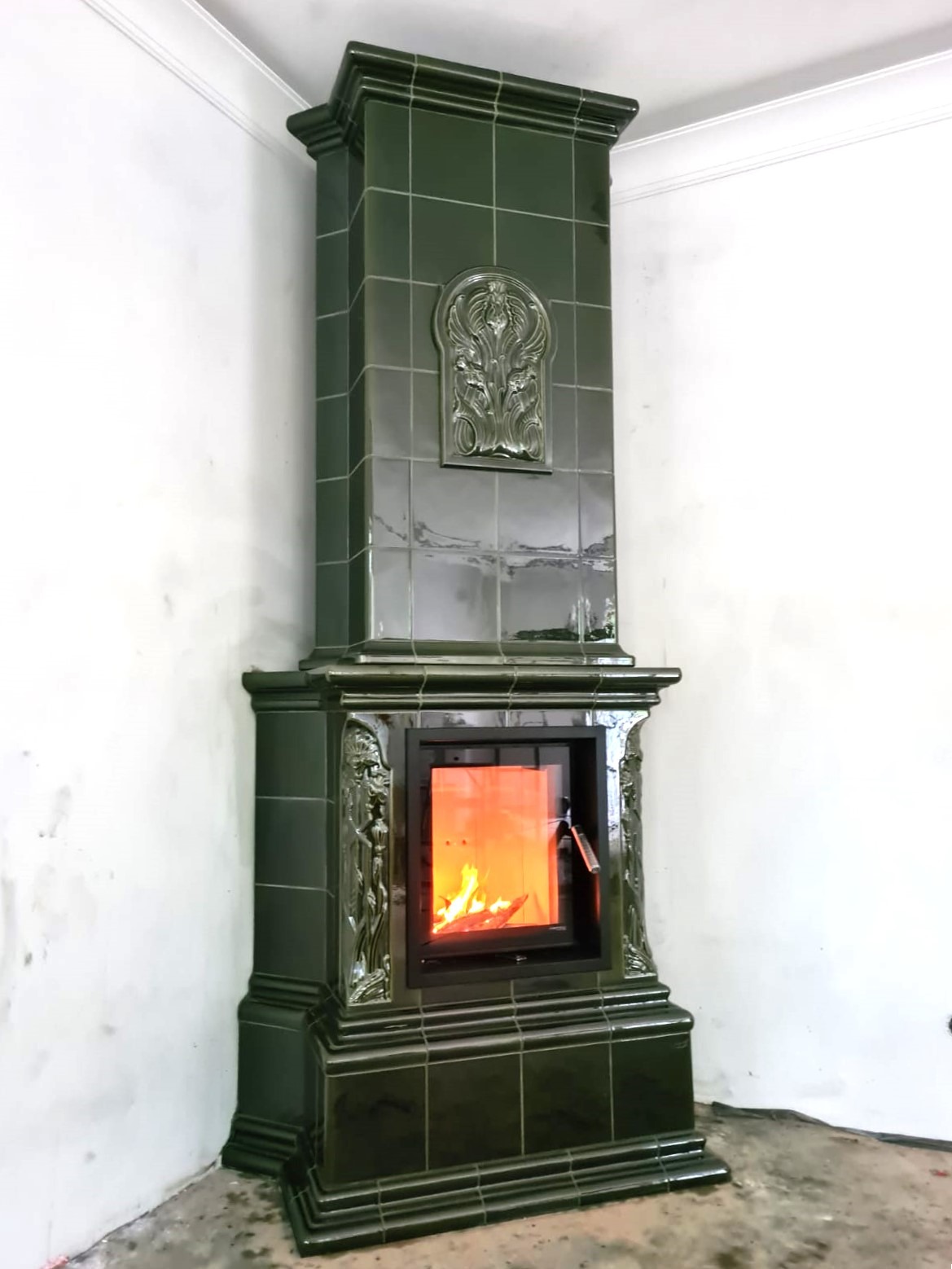 Zdobiony, zielony kominek kaflowy z palącym się ogniem, umieszczony w rogu pomieszczenia z białymi ścianami. Widoczne detale zdobień i faktura kafli.