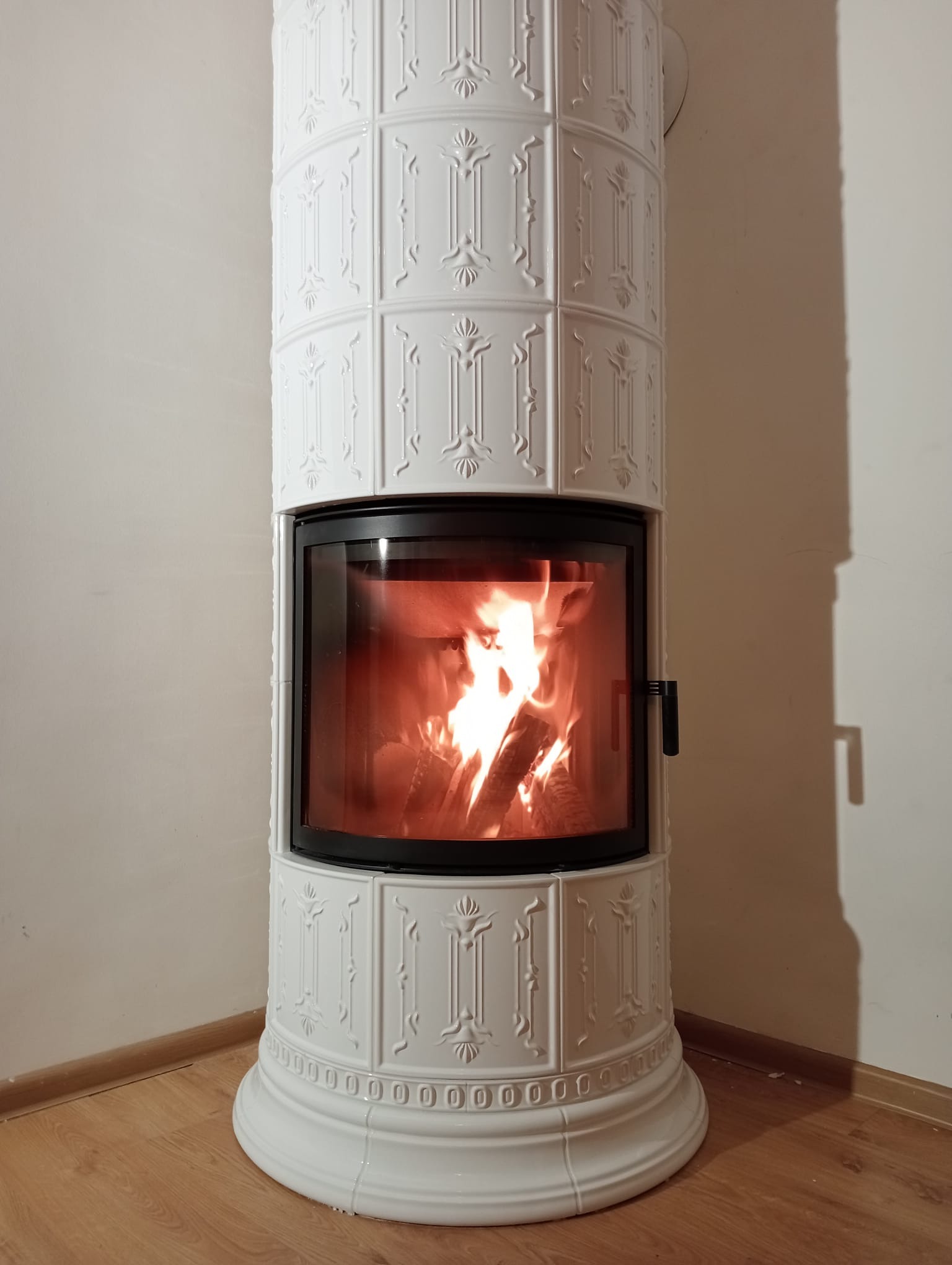 Biały, kaflowy kominek z palącym się drewnem, widoczny w przeszklonym palenisku. Dekoracyjne zdobienia na kaflach, ciepłe światło ognia. Ujęcie z bliska, w rogu pomieszczenia.