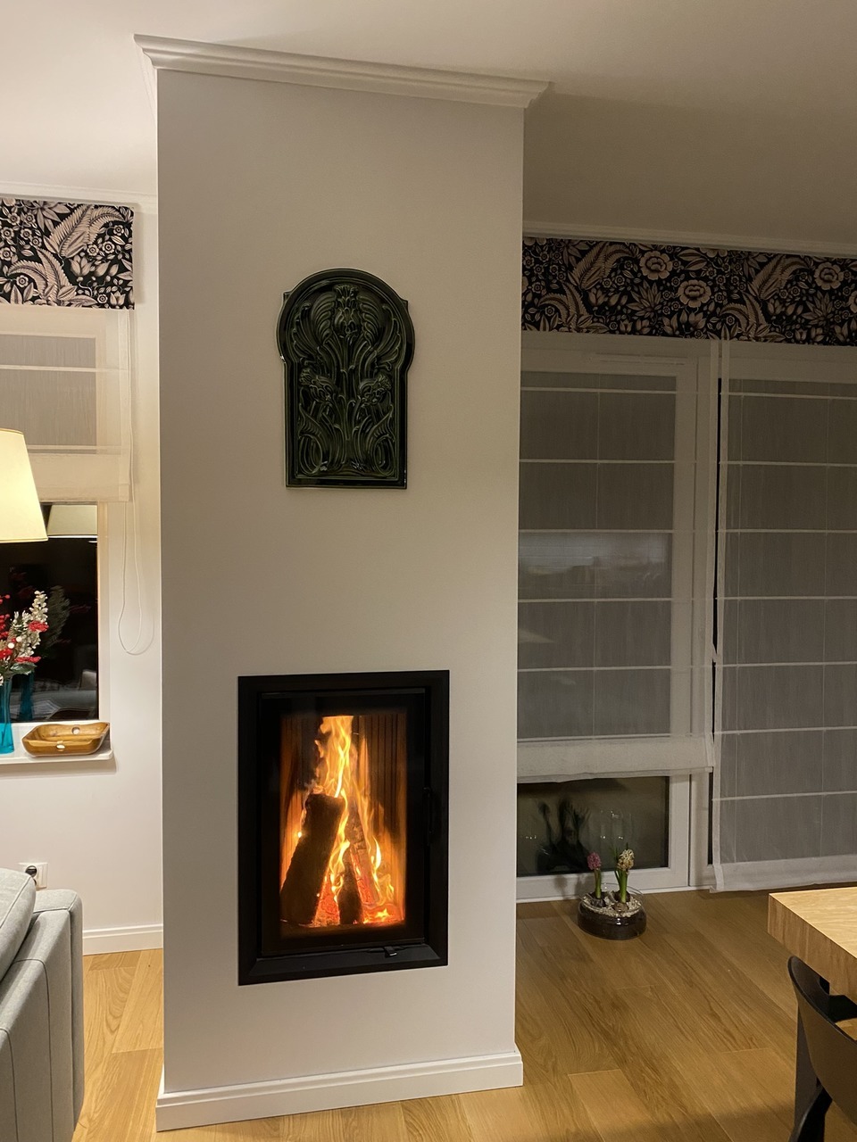 Nowoczesny kominek w zabudowie z ozdobną płytką ceramiczną nad paleniskiem. Widoczne płomienie i drewno wewnątrz. W tle okno z roletami rzymskimi i dekoracyjny wazon.