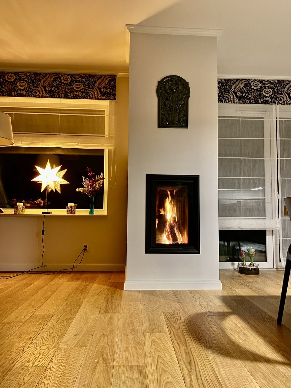 Nowoczesny kominek z czarną obudową i płomieniami, ozdobna płaskorzeźba nad nim, drewniana podłoga, dekoracje okienne w kwiatowe wzory, ciepłe oświetlenie.