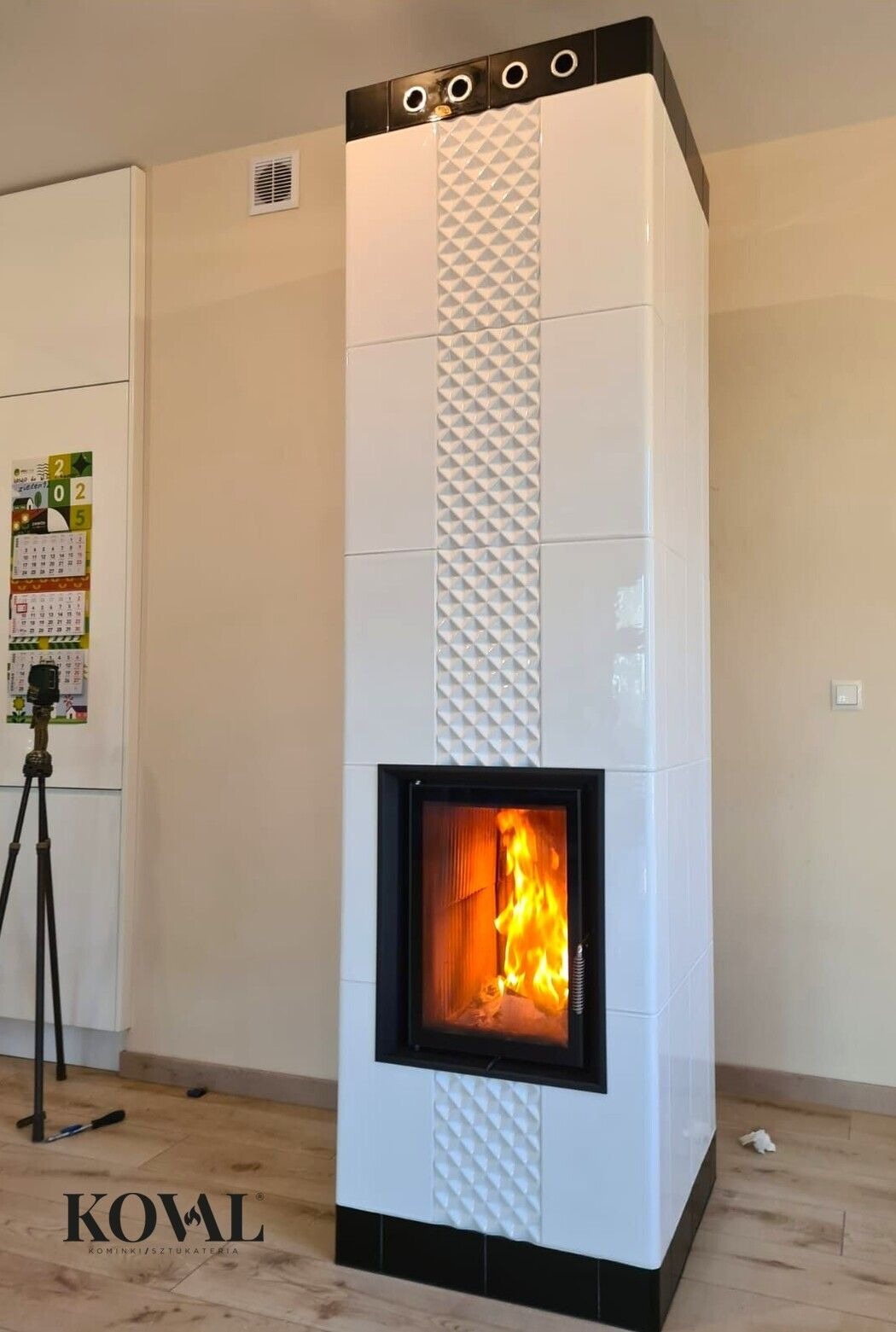Biały, nowoczesny kominek kaflowy z czarnymi akcentami, płomienie widoczne przez szybę, dekoracyjny wzór 3D na kaflach, wnętrze w neutralnych barwach.