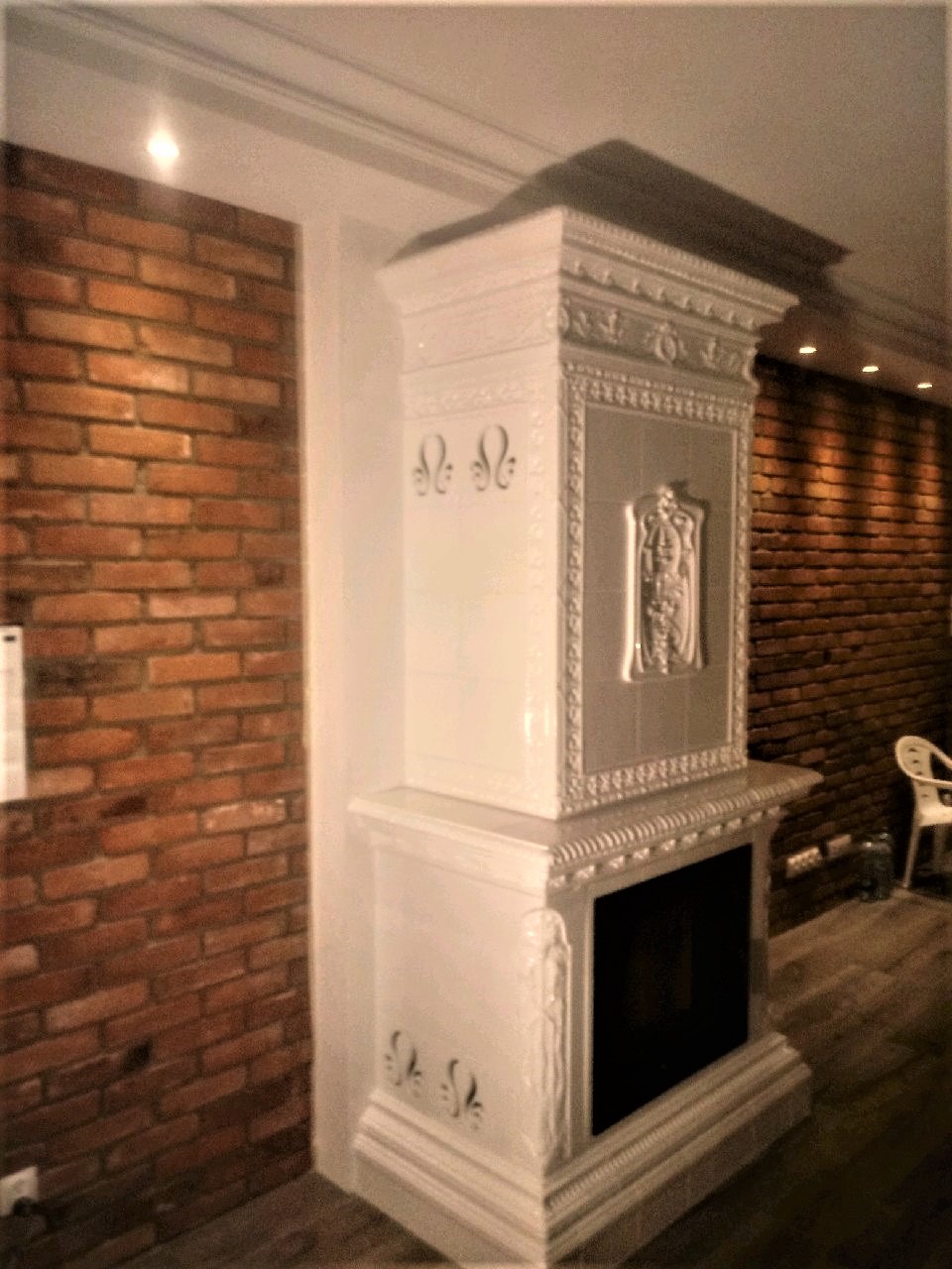 Biały, ozdobny piec kaflowy z czarnym paleniskiem, ustawiony w rogu pomieszczenia z ceglanymi ścianami, oświetlony punktowo.