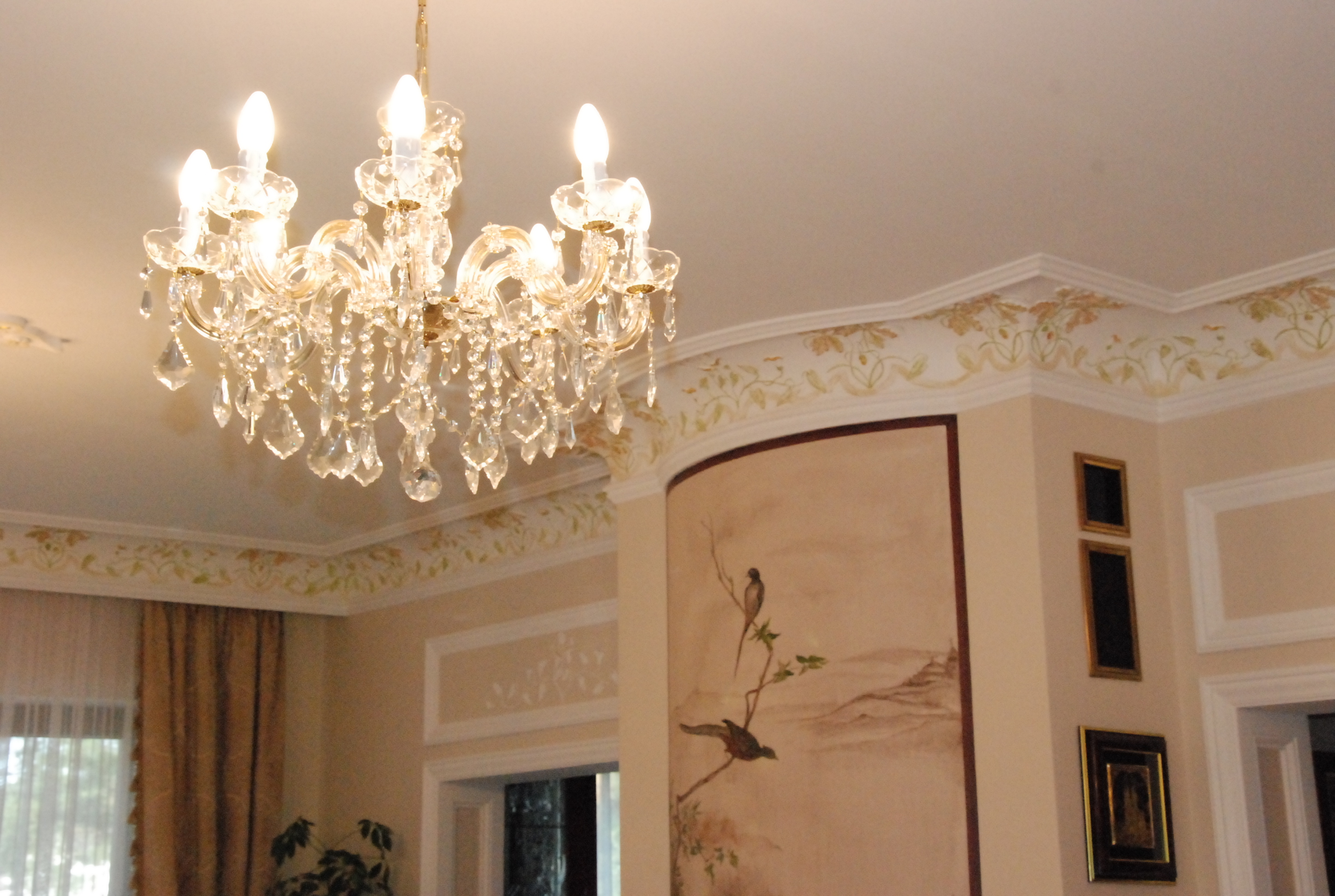 Elegancki, kryształowy żyrandol oświetla jasne wnętrze z dekoracyjnym malowaniem na suficie i ścianie, przedstawiającym motywy roślinne i ptasie.