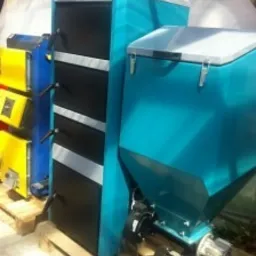 Kocioł na paliwo stałe z podajnikiem ślimakowym, marki Radom, w kolorze turkusowo-czarnym, na palecie. Widoczne również fragmenty innych kotłów w tle.