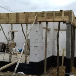 Wznoszenie ścian domu z bloczków betonowych, z widocznym deskowaniem stropu i podporami z gałęzi drzew, na fundamencie zabezpieczonym hydroizolacją.