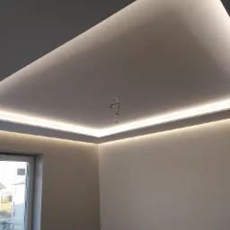 Nowoczesny sufit podwieszany z oświetleniem LED w narożach, widoczna żarówka na kablu, okno z widokiem na budynek w tle.