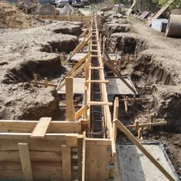 Szeroki kadr na wykop pod fundamenty z drewnianym szalunkiem i zbrojeniem stalowym, widoczne nierówności terenu i przygotowany grunt pod wylewkę.