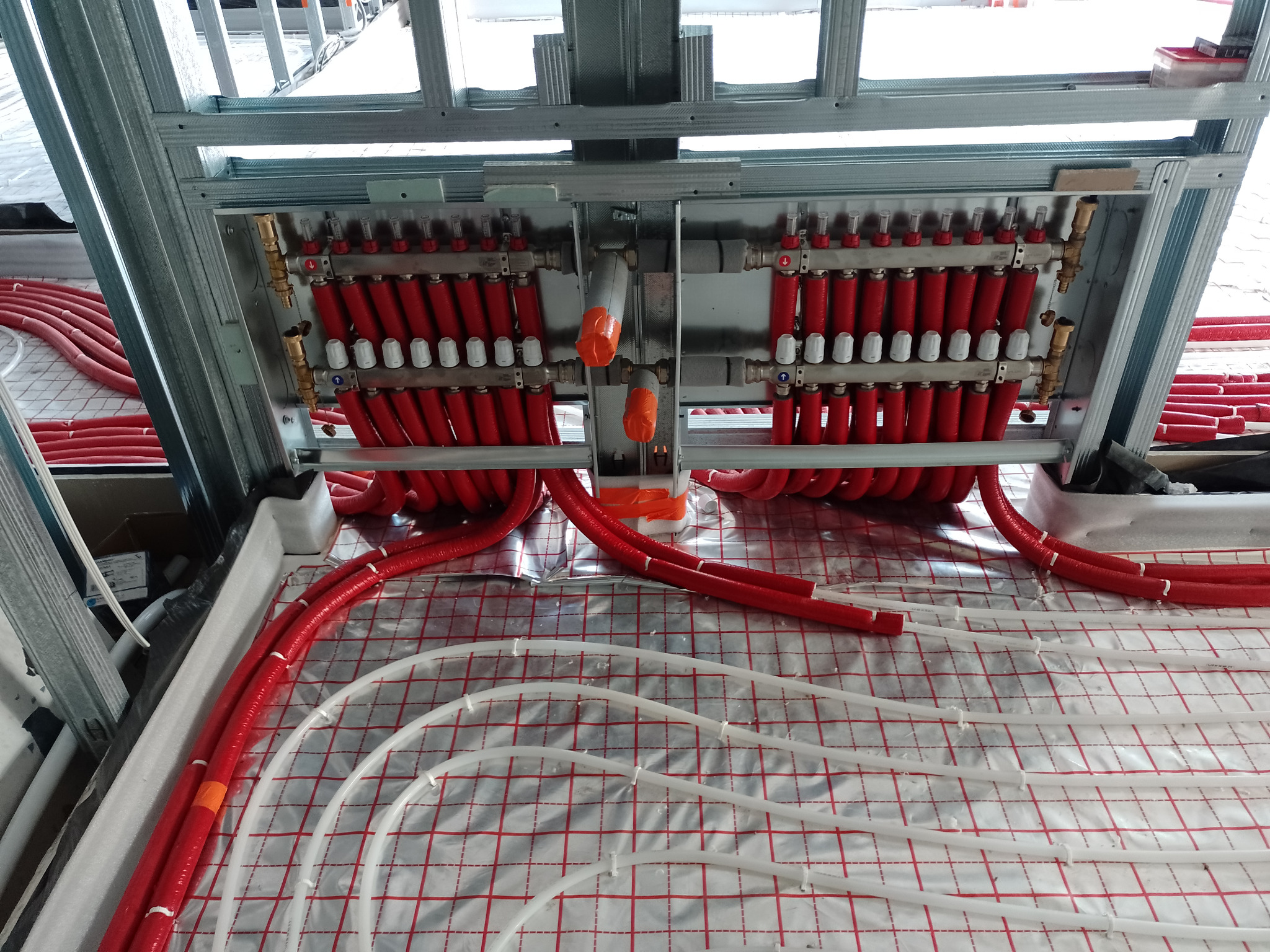 Rozdzielacz ogrzewania podłogowego z czerwonymi rurami, zamontowany na metalowej konstrukcji, widoczne białe rurki na folii z czerwoną siatką.