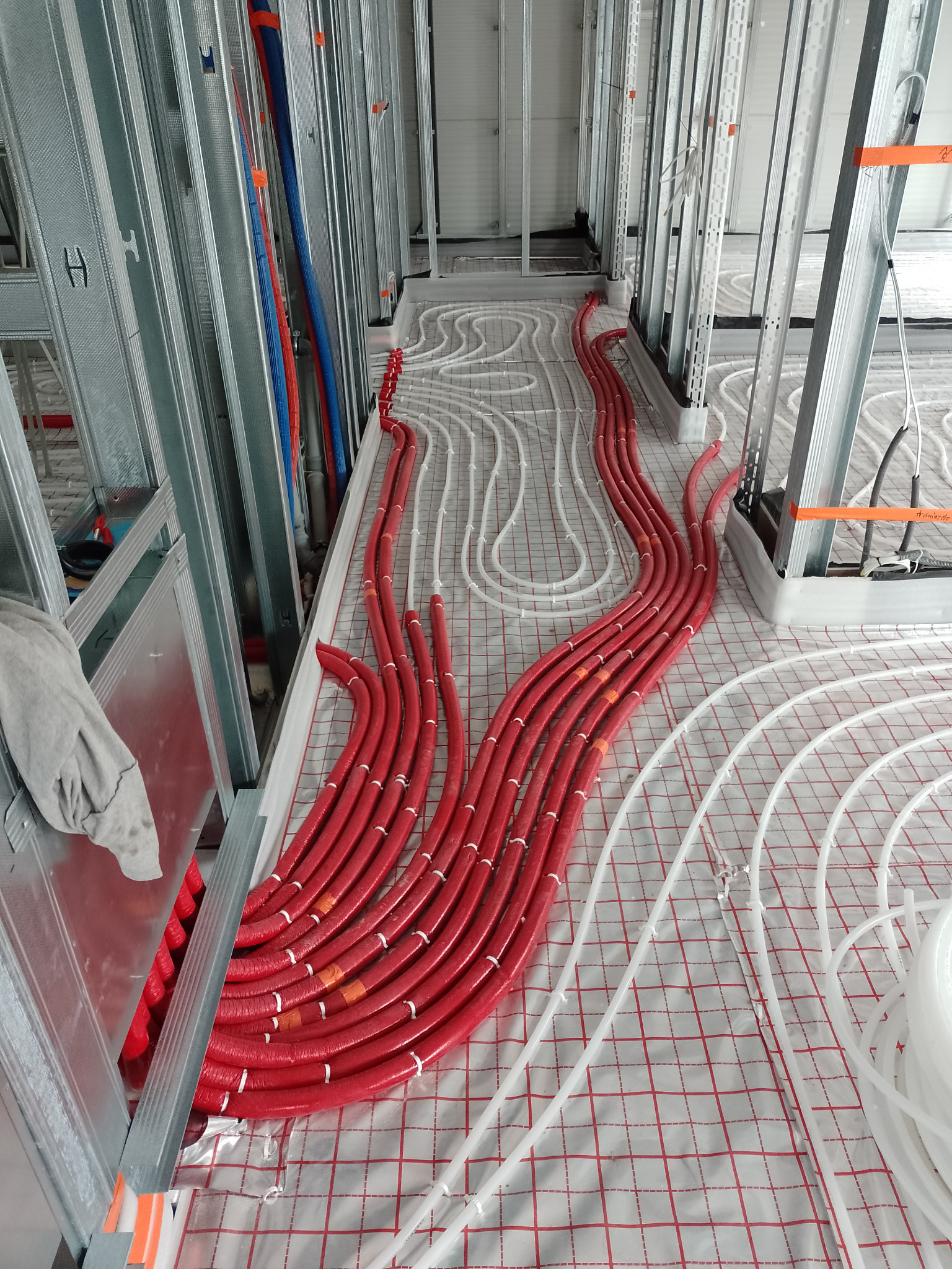 Instalacja ogrzewania podłogowego z czerwonych i białych rur na folii z siatką, widoczne metalowe profile ścian działowych.