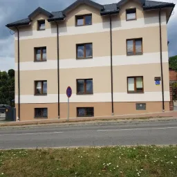 Trzykondygnacyjny budynek z elewacją w odcieniach beżu i brązu, z poziomymi pasami i ciemnymi oknami, widoczny z poziomu ulicy, z zielenią i znakami drogowymi w tle.