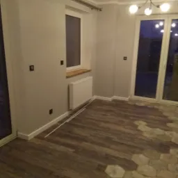 Pokój po remoncie z panelami podłogowymi imitującymi drewno, fragmentem podłogi wyłożonym heksagonalnymi płytkami i świeżo pomalowanymi ścianami w odcieniach szarości.