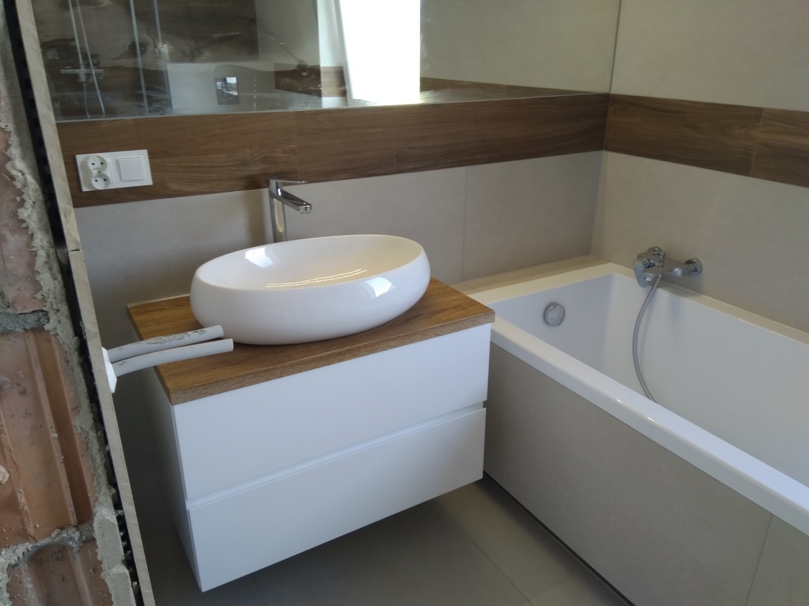 Nowoczesna łazienka z owalną umywalką nablatową na szafce z drewnianym blatem, obok wanna obudowana płytkami w odcieniach beżu, widoczne rury wystające ze ściany przed wykończeniem.
