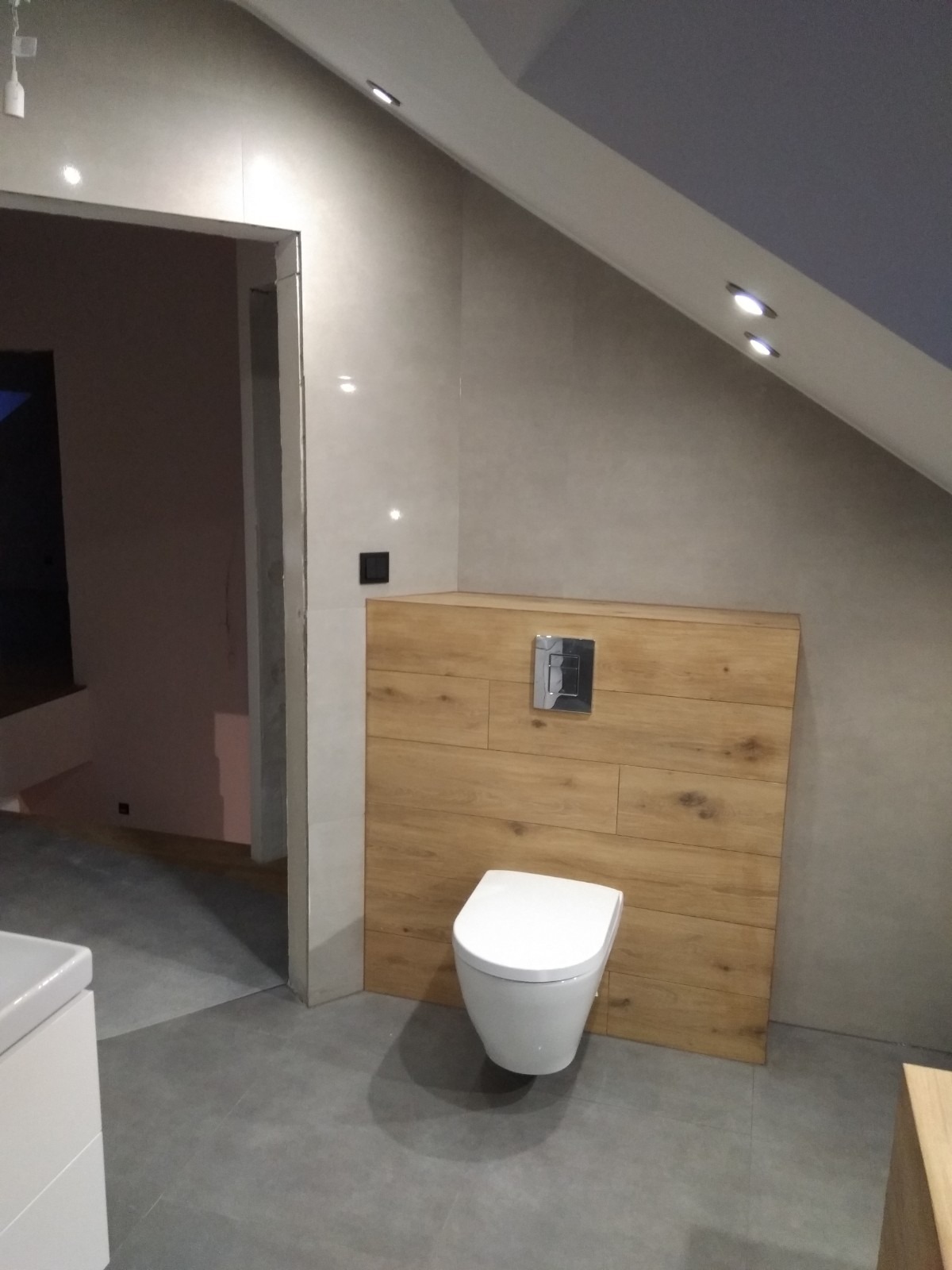 Nowoczesna łazienka na poddaszu ze stelażem podtynkowym obudowanym drewnopodobnymi panelami, wisząca toaleta i szare płytki podłogowe.