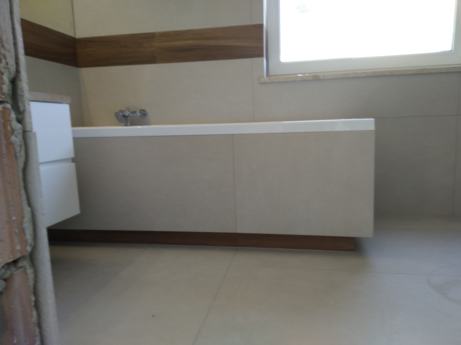 Nowoczesna łazienka w trakcie wykańczania: widoczna zabudowana wanna z beżowymi płytkami, biała szafka umywalkowa i fragment surowej ściany w lewym dolnym rogu.