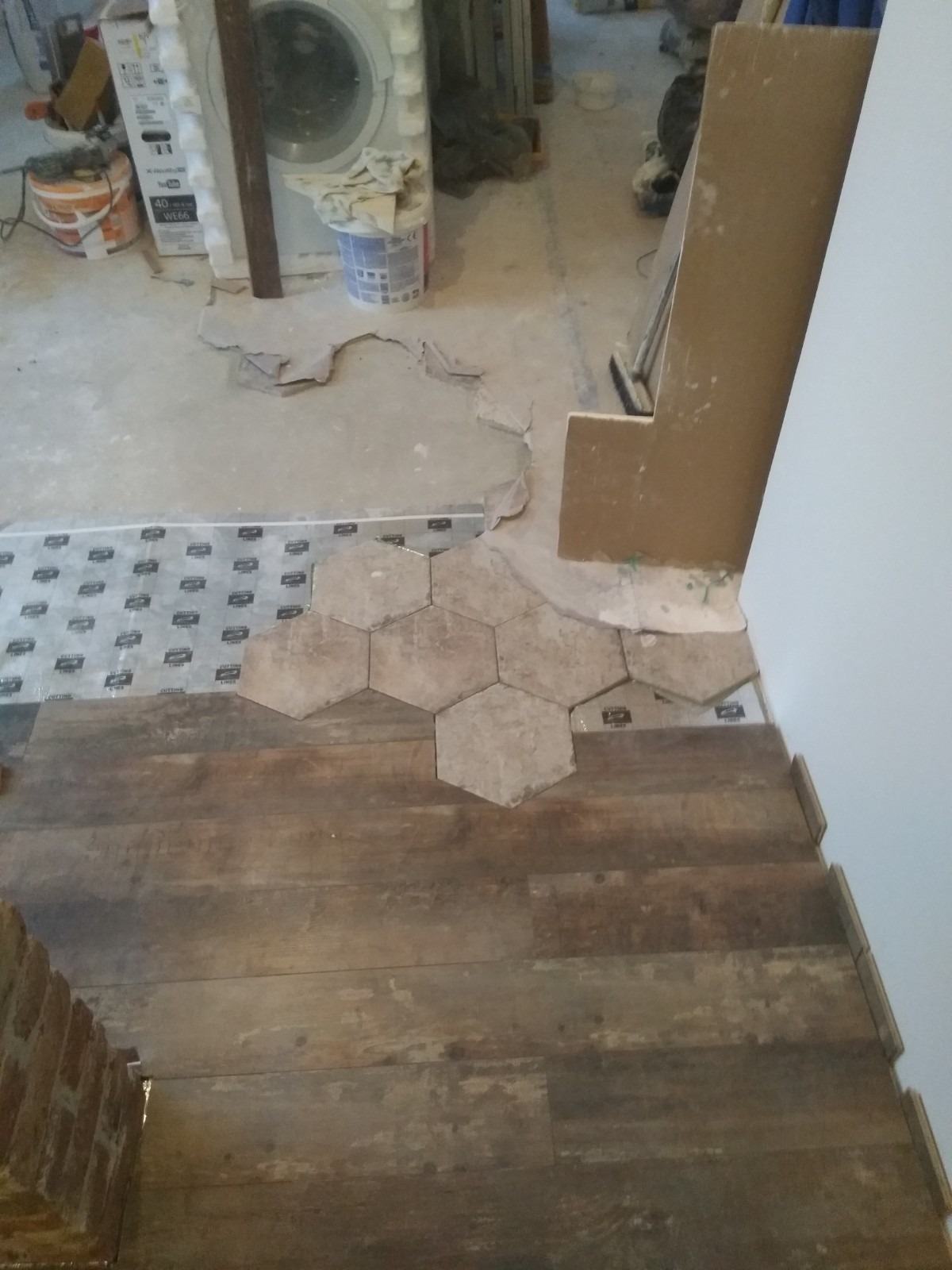 Fragment podłogi w trakcie łączenia drewnopodobnych paneli z heksagonalnymi płytkami, widoczne podłoże z folią izolacyjną i pozostałości zaprawy, w tle pralka i narzędzia.