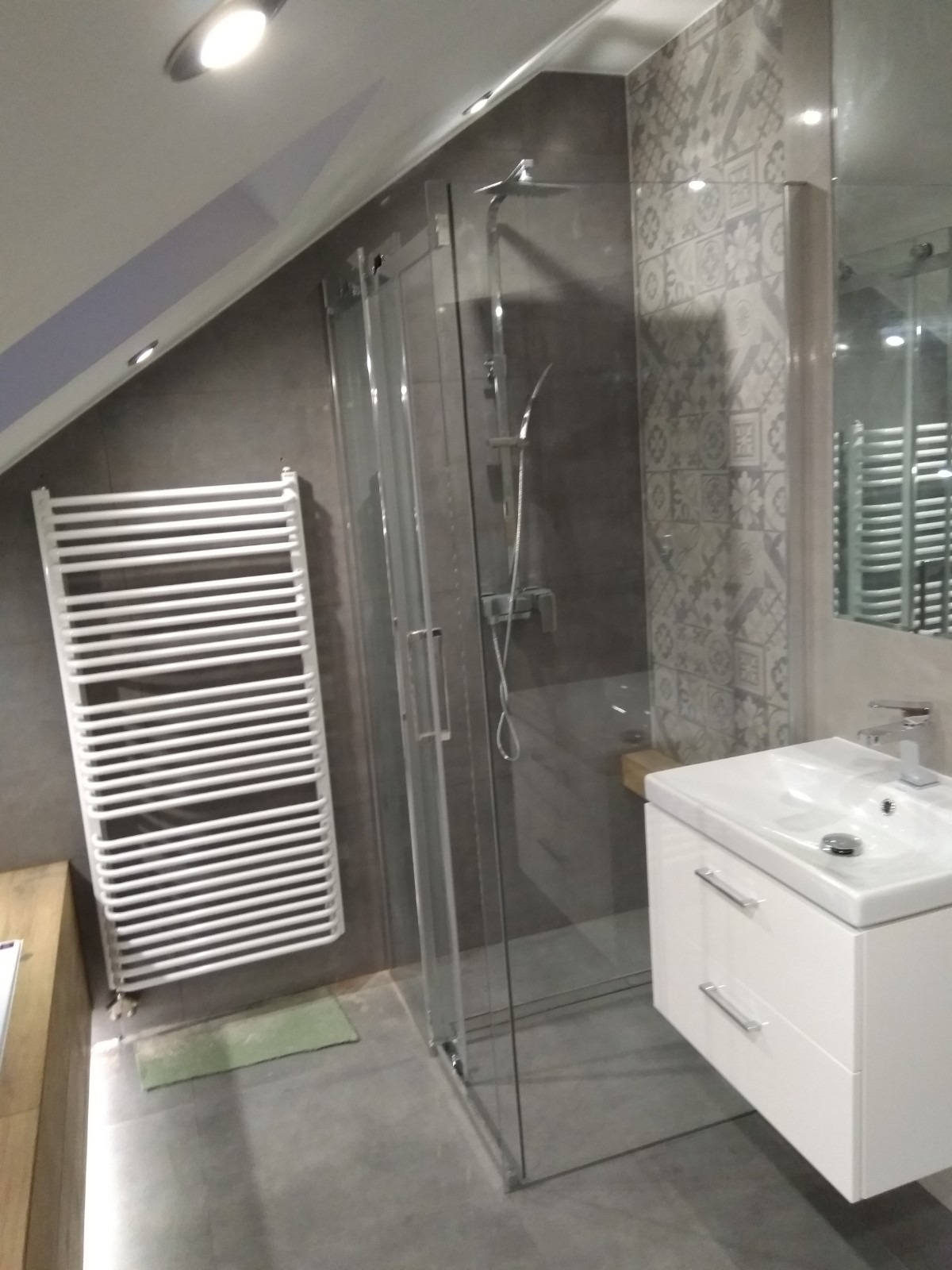 Nowoczesna łazienka na poddaszu z kabiną prysznicową ze szklanymi drzwiami przesuwnymi, białą umywalką z szafką, grzejnikiem drabinkowym i szarą płytką dekoracyjną na ścianie.