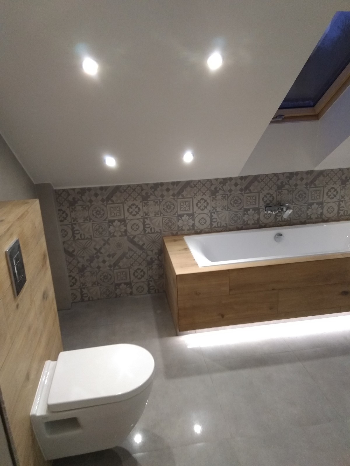 Nowoczesna łazienka na poddaszu z białą wiszącą toaletą, zabudowaną wanną z drewnianym wykończeniem i podświetleniem LED oraz wzorzystymi płytkami na ścianie.