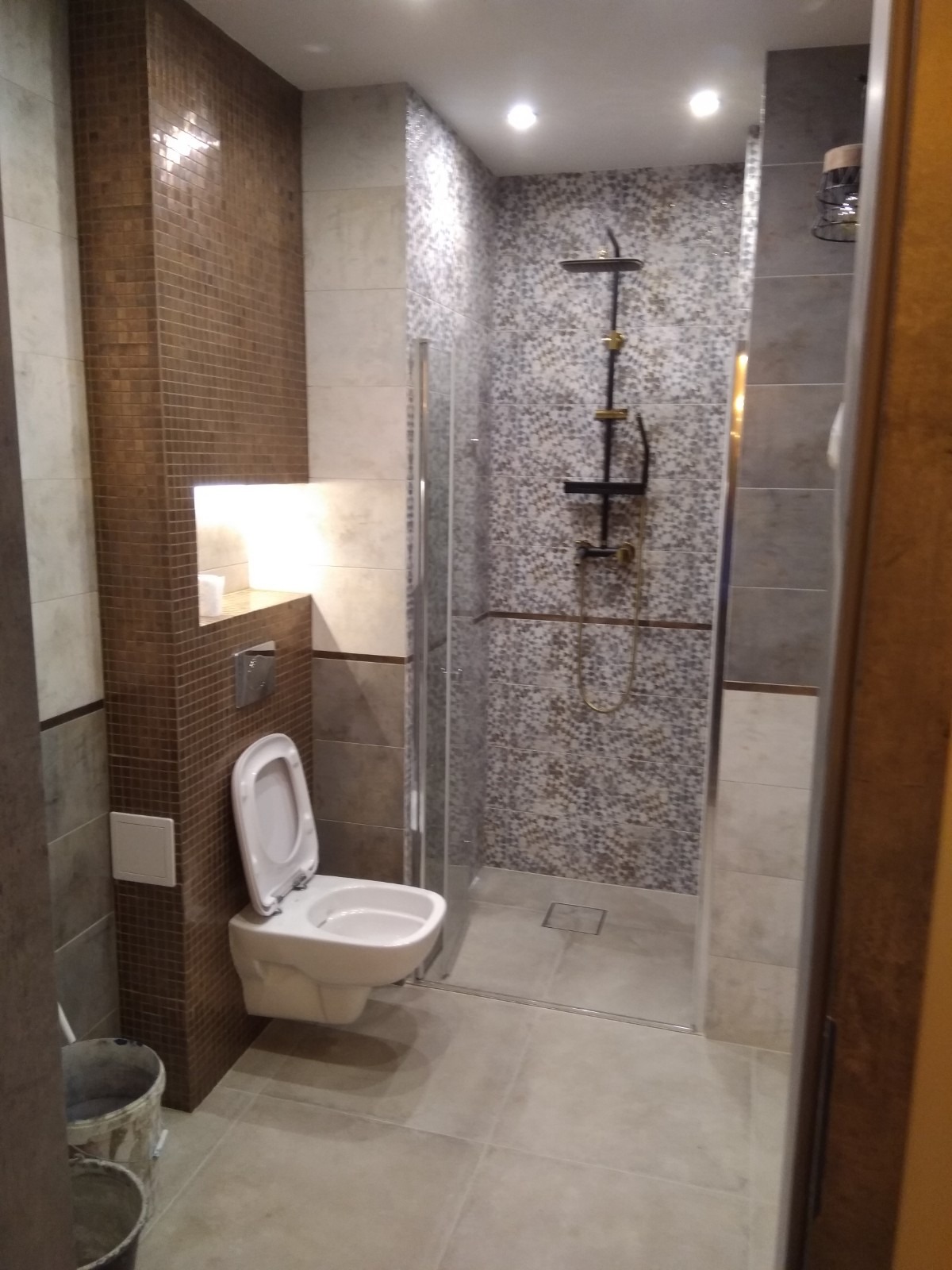 Nowoczesna łazienka po remoncie: kabina prysznicowa typu walk-in z deszczownicą w kolorze czarnym i złotym, toaleta podwieszana oraz ściana wyłożona brązową mozaiką z wnęką oświetloną LED.