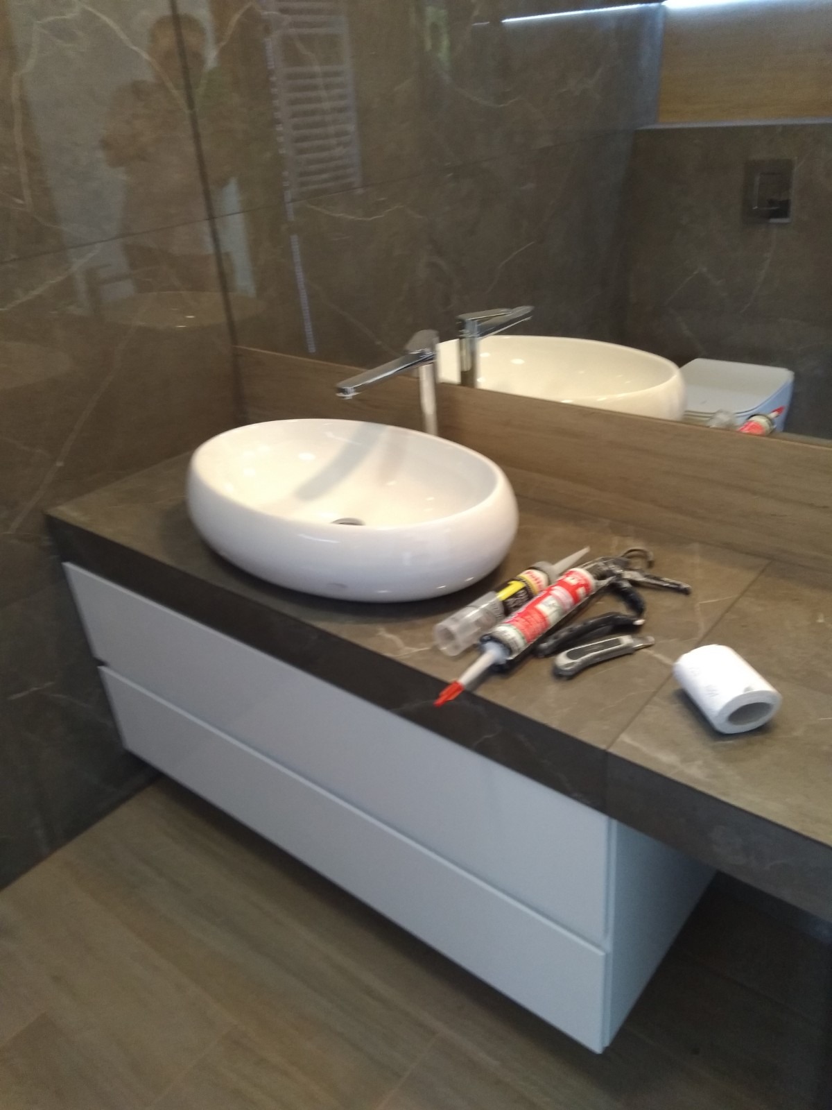 Owalna umywalka nablatowa z chromowaną baterią na blacie z ciemnego marmuru. Widoczne dwie tuby silikonu, nożyk i rolka papieru toaletowego. W tle lustro i brązowe płytki.