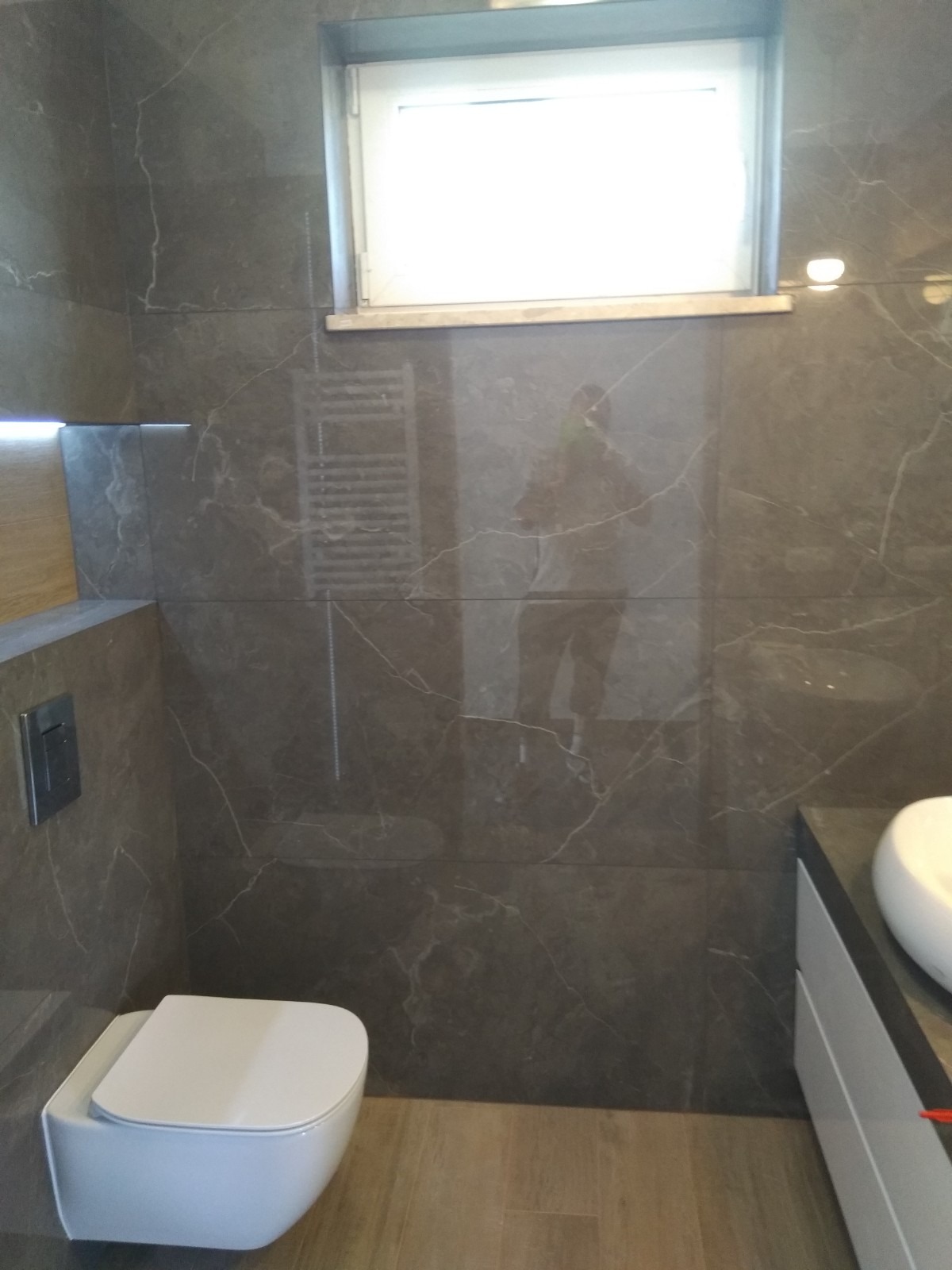 Nowoczesna łazienka z szarymi płytkami imitującymi marmur, podwieszana toaleta i grzejnik drabinkowy widoczny w odbiciu na ścianie.