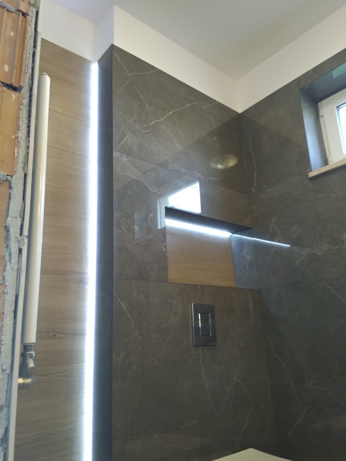 Fragment łazienki w trakcie wykańczania: widoczne płytki imitujące marmur, podświetlenie LED wzdłuż ściany, częściowo obudowany stelaż WC z przyciskiem spłukującym i niedokończone wykończenie...