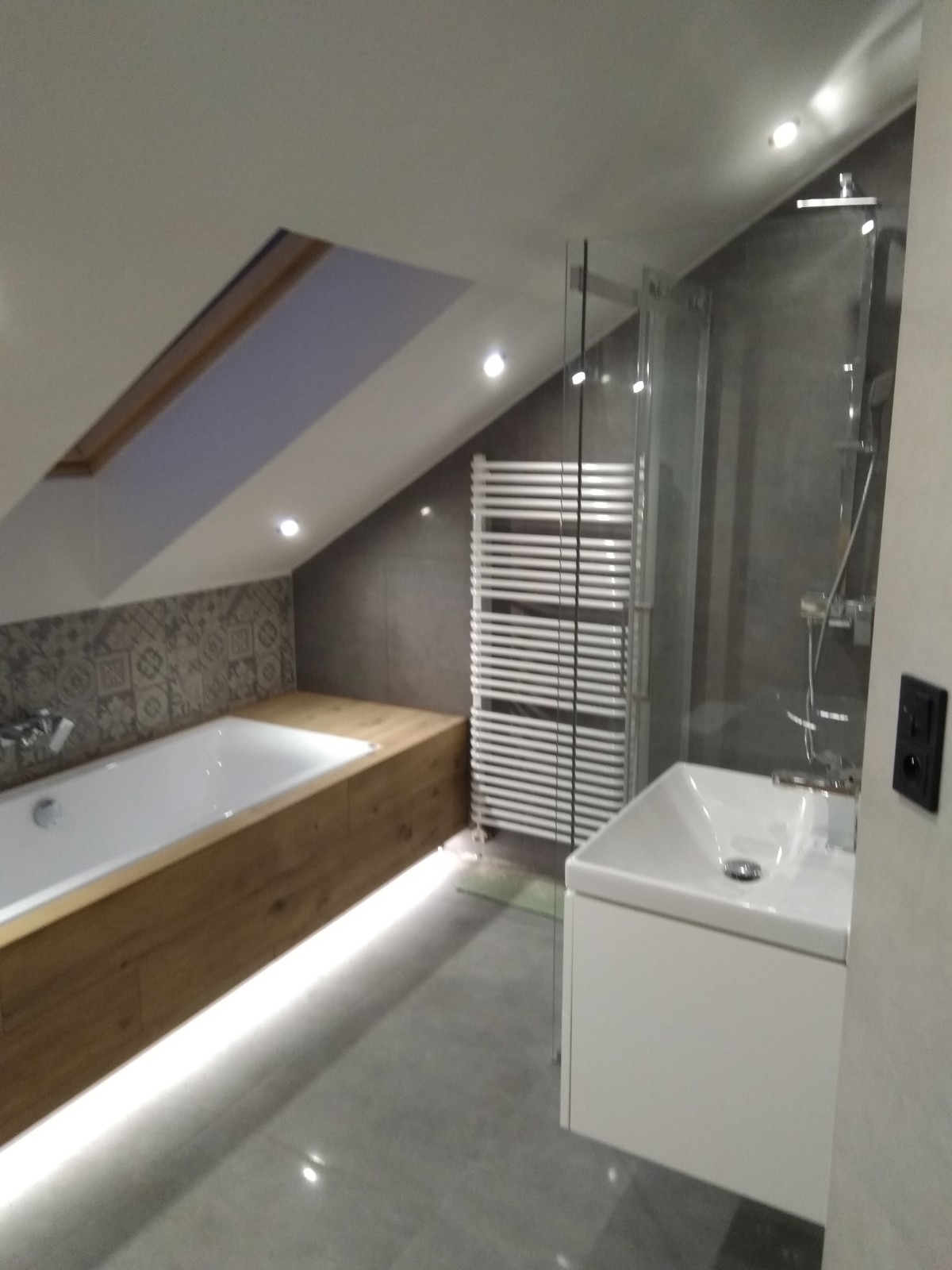 Nowoczesna łazienka na poddaszu z wanną obudowaną drewnem, podświetleniem LED, grzejnikiem drabinkowym i szklaną kabiną prysznicową.