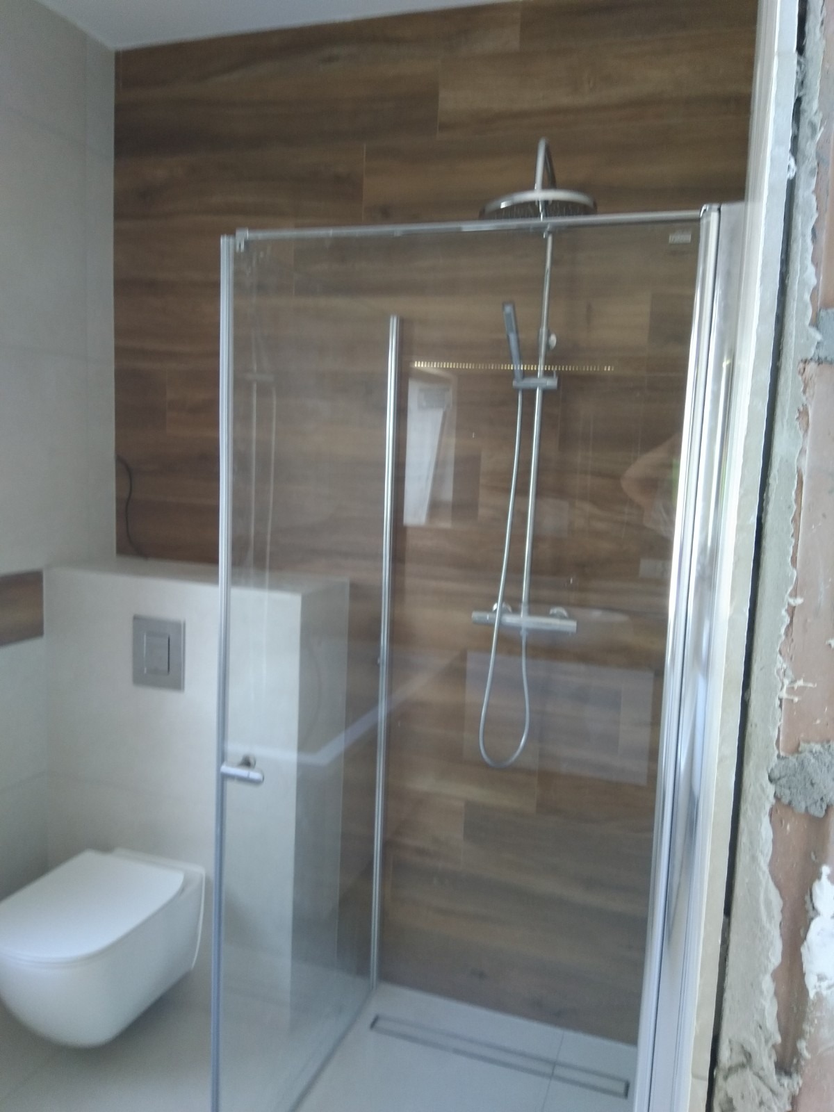 Nowoczesna kabina prysznicowa ze szklanymi drzwiami, deszczownicą i drewnopodobnymi płytkami na ścianie, widoczny fragment toalety podwieszanej i niedokończone wykończenie ściany po prawej stronie.